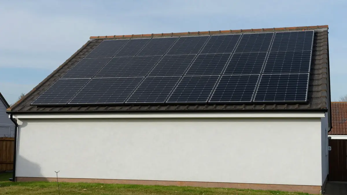 A pitched tiled roof on a white-rendered UK house with a large array of black solar panels, seen side-on from the garden under a blue sky