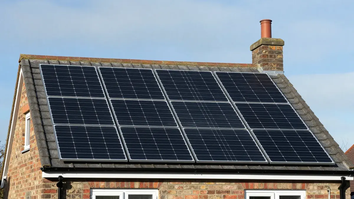A grey-tiled roof on a brick house covered by a neat rectangular array of black solar panels, with a chimney stack and blue sky behind