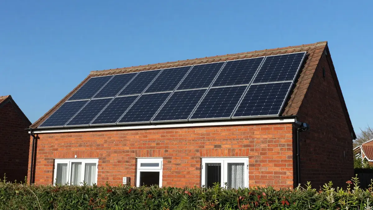 A red-brick UK house with two rows of black solar panels on the roof in bright sunshine, a green hedge in front and a deep blue sky